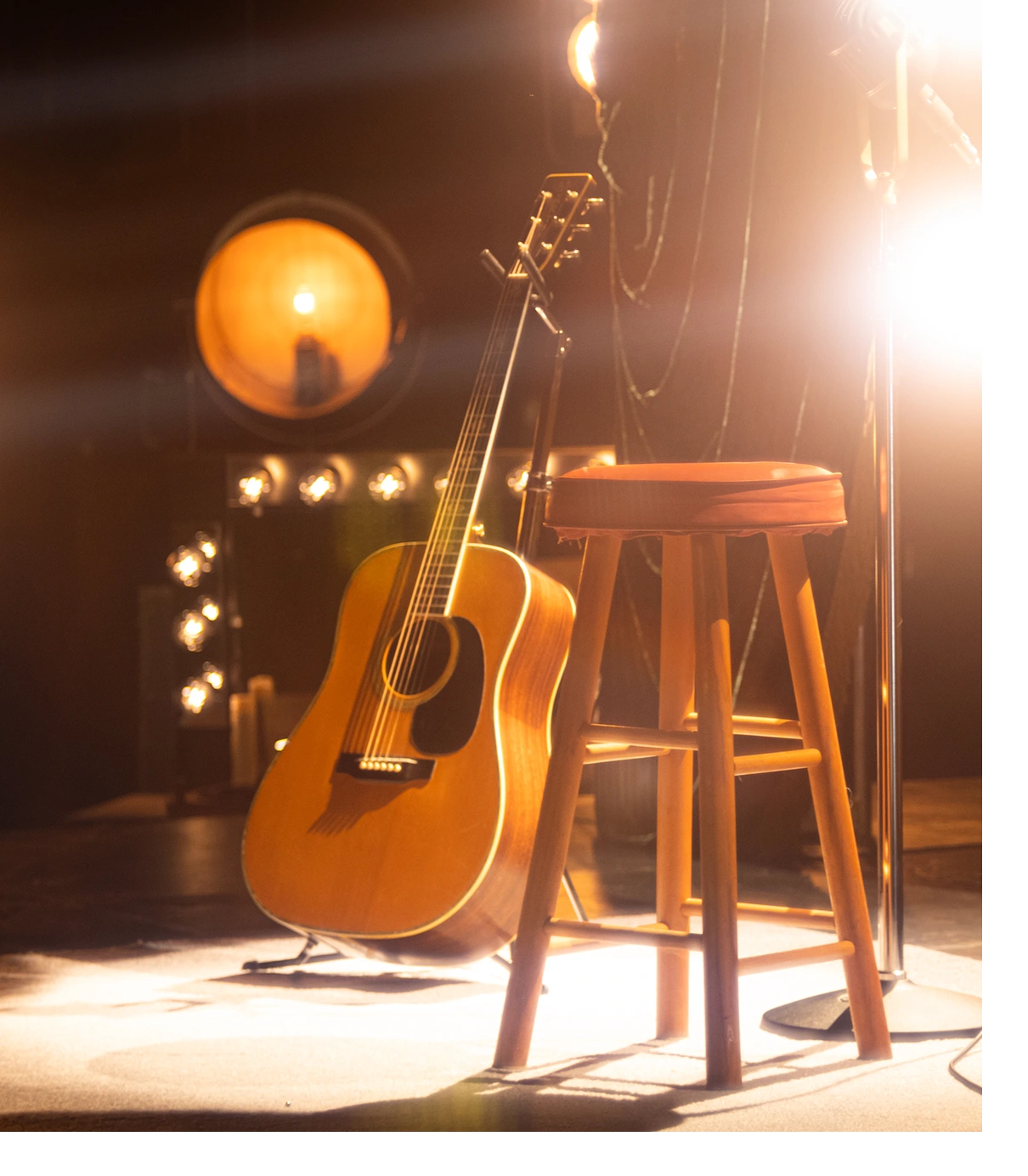 Acoustic guitar resting against a wooden stool on a warmly lit stage.